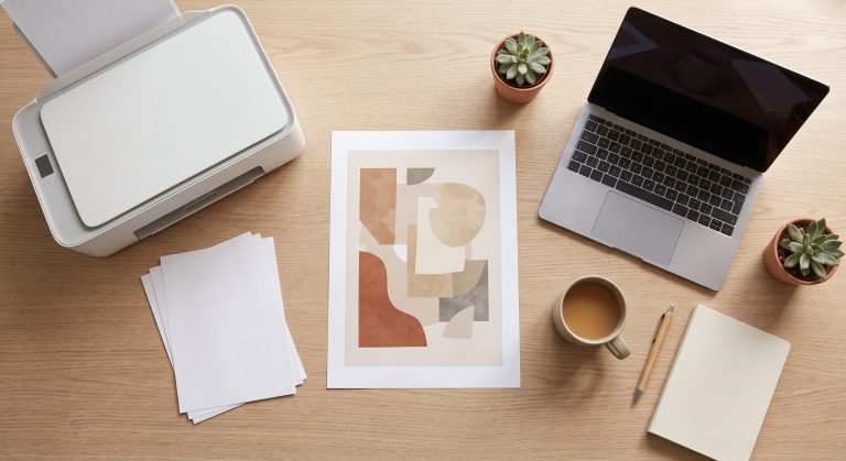 Flat lay of a printed abstract artwork sheet on a desk beside a laptop and a home printer